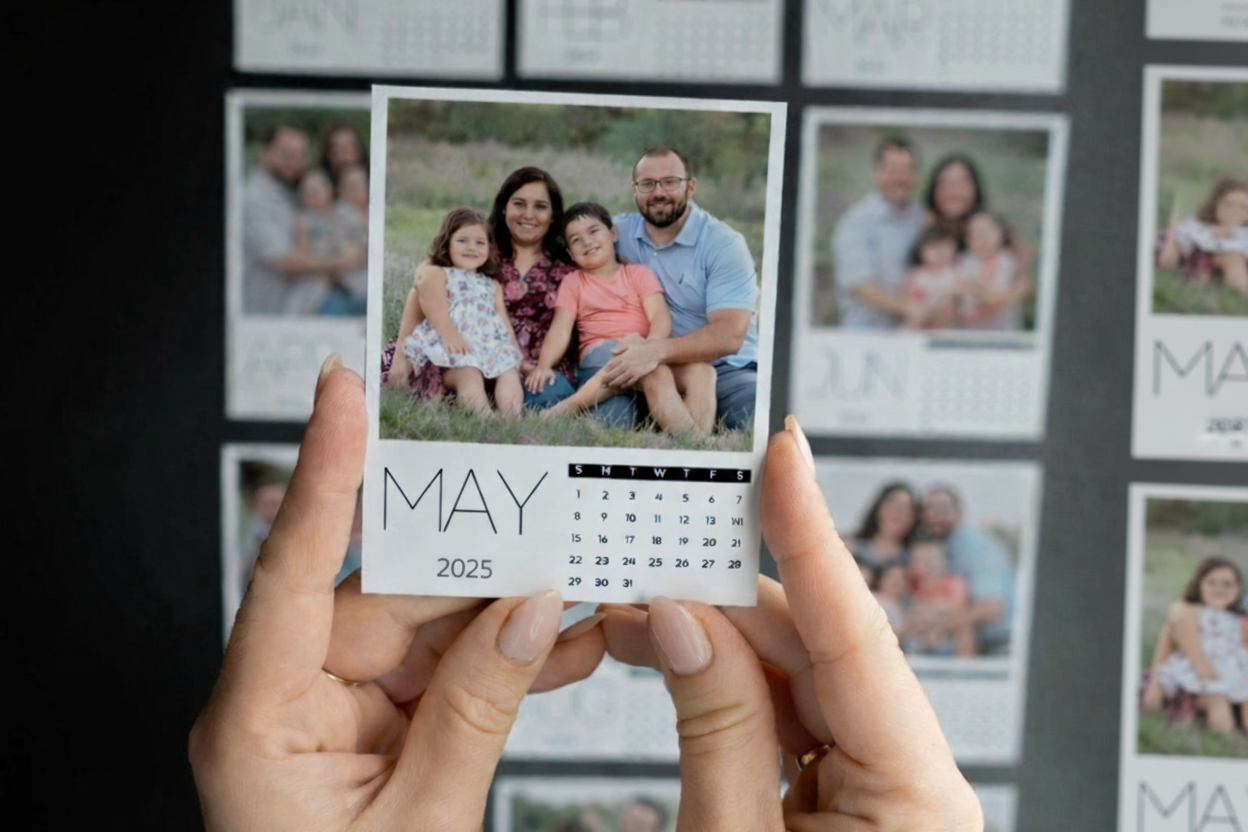 Hand holding a May 2025 calendar with family photos displayed on a wall.