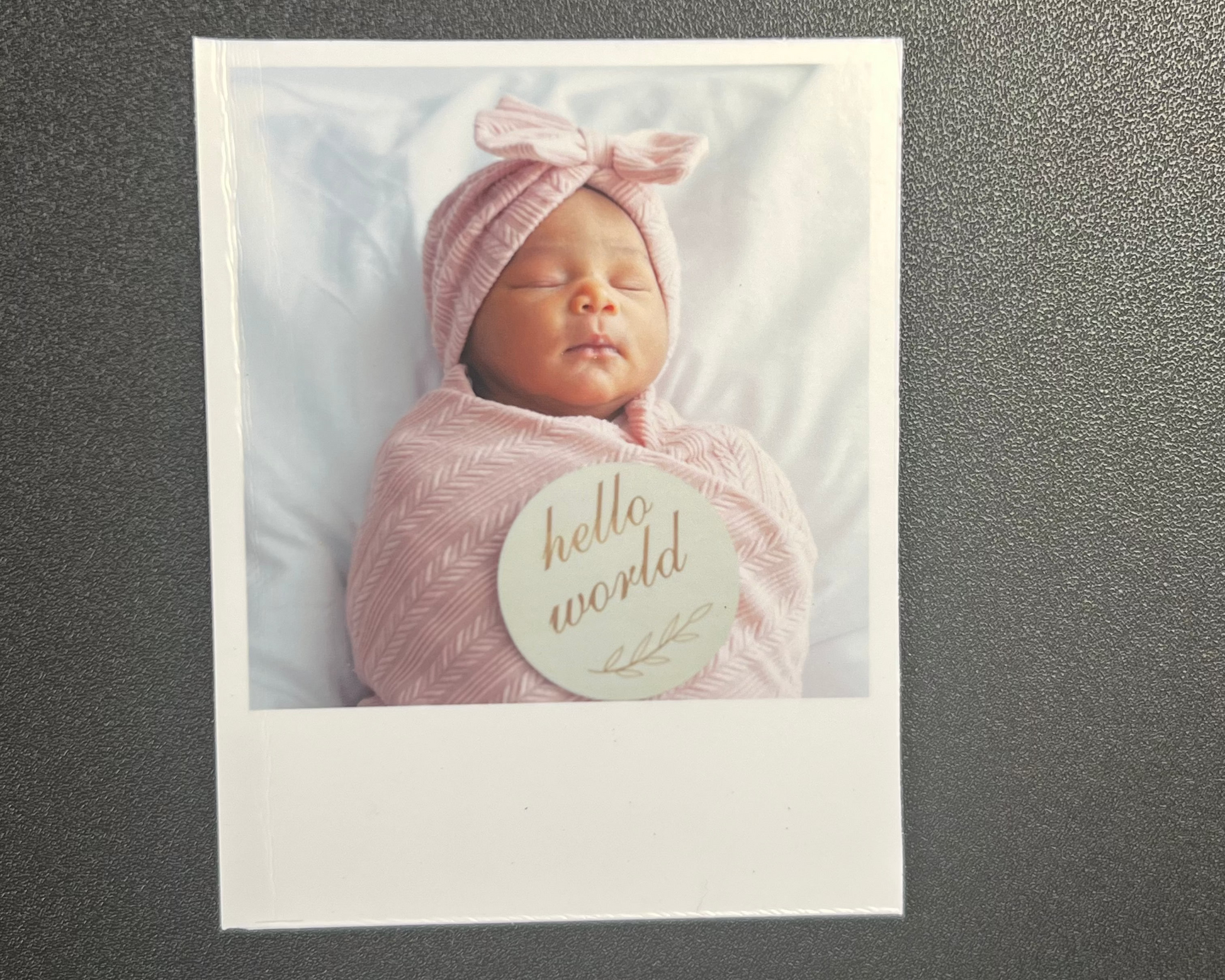 Newborn baby wrapped in pink with a 'hello world' card on a dark surface